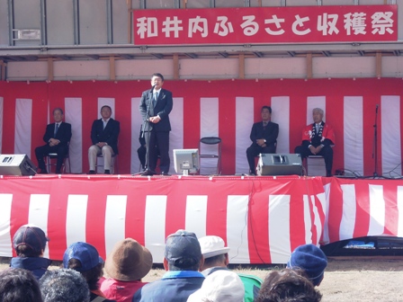 「和井内ふるさと収穫祭」と書かれた吊り看板に紅白幕がはられた野外ステージ上に、関係者が椅子に座り、ステージ中央でマイクスタンドの前に立つ市長の写真