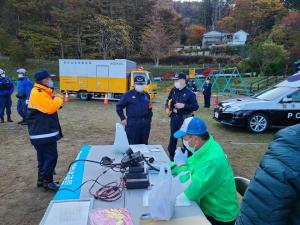 白と黄色のツートンのトラックやパトカーが駐車した遊具が設置された公園内で、緑の蛍光色のスタッフジャンパーを着て青色のキャップを被った男性が、テーブルの上に設置された器機から伸びたマイクを持ち放送を行っている様子を、紺色の作業着を着用した市長が見ている防災訓練の様子の写真