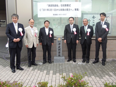 ”「津波到達地」石柱除幕式（「2011年3月11日から未来の貴方へ」事業）”と書かれた紙が大きな窓ガラスに貼られ、その前に建てられた石柱の両隣りに3名ずつスーツを着た男性達が並び記念撮影している写真
