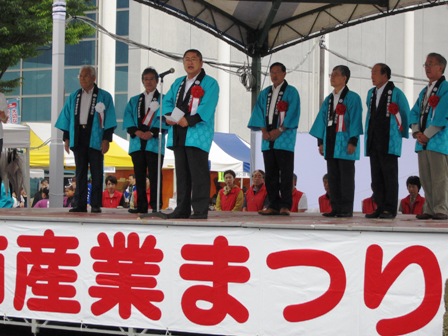 野外ステージ後方で、水色のはっぴを着て胸に胸章リボンを付けた関係者が立っており、ステージ中央に設置されたマイクスタンドの前に立つ市長が挨拶を行っている写真