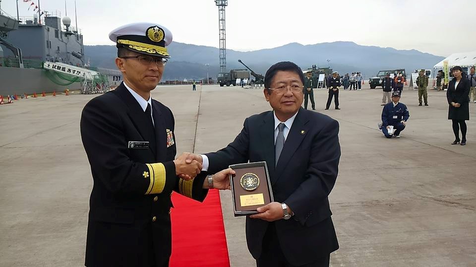 制服を着た海上自衛隊試験艦「あすか」の乗組員の方と市長が盾を右手で一緒に持ち、左手で握手をしている様子の記念写真