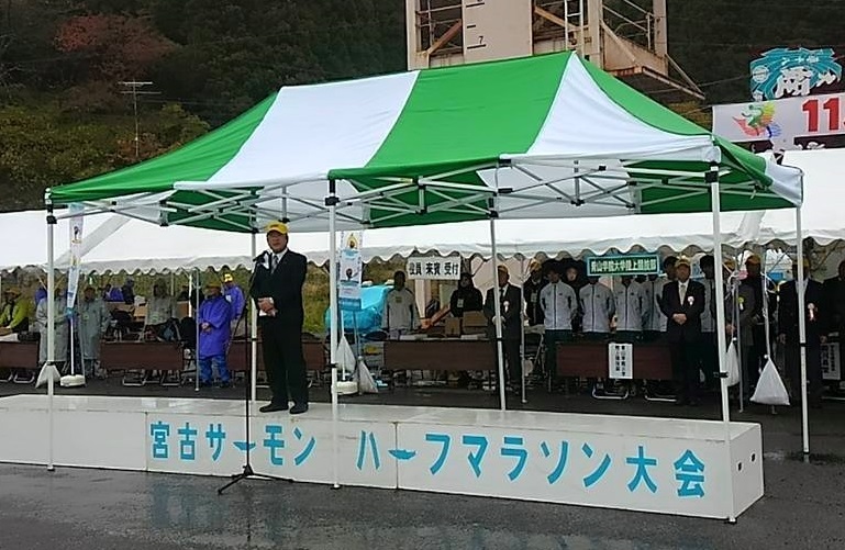 緑色と白色のテントの下で、「宮古サーモンハーフマラソン大会」と書かれた台の上に立ちスタンドマイクに向かい話をする市長の写真