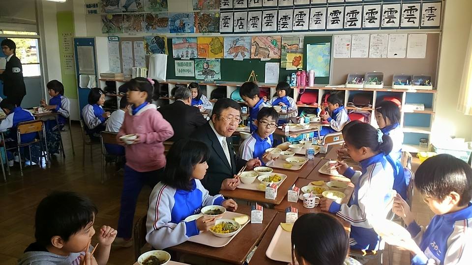教室で向かい合わせにした机の席で給食を食べる市長と小学生たちの写真