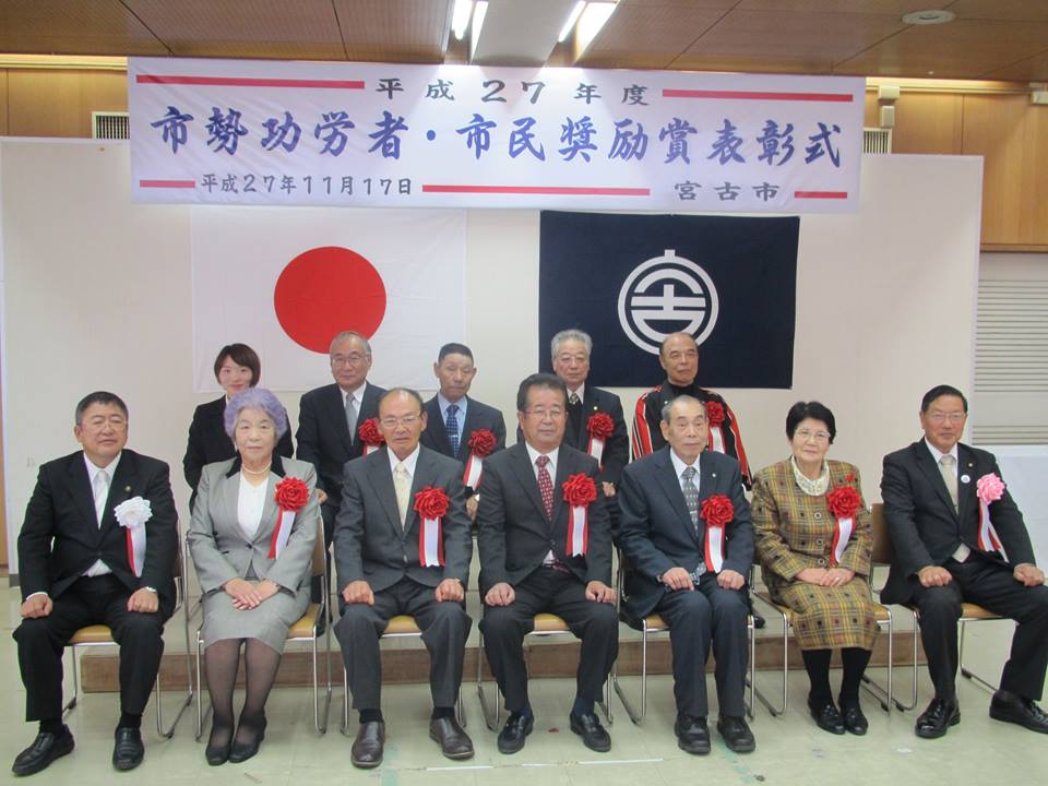 「平成27年度 市勢功労者・市民奨励賞表彰式」と書かれた横断幕の下で、椅子に座る市長と11名の受賞者たちが2列に並んで写る記念写真