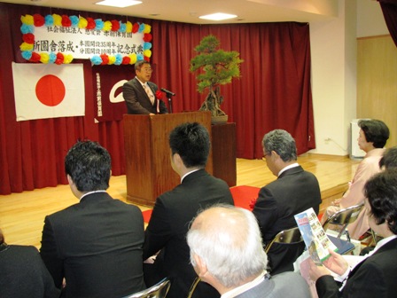 カラフルなペーパーフラワーで囲まれた吊り看板が設置されたステージ中央で、演壇に立つ市長と、新園舎落成式に参列する参加者の写真