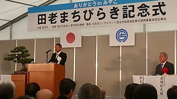 「田老まちびらき記念式」と書かれた横断幕、日本国旗、宮古市の市町村旗が掲げられた舞台上で、演台に置かれたスタンドマイクに向かい話をする市長の写真