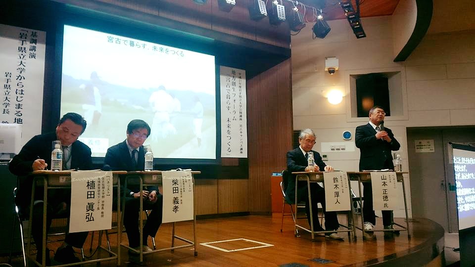 画面が映されたスクリーンが設置された舞台で、左右に置かれた机の席に座る3名の男性と、立って話す市長の写真