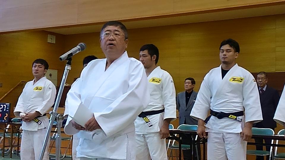 柔道着を着て横一列に並ぶ男性たちの前で、柔道着を着てスタンドマイクに向かい話をする市長の写真