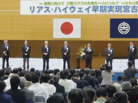 体育館に大勢の参加者が椅子に座っており、「リアス・ハイウェイ早期実現宮古大会」と書かれた横断幕の下、来賓席前で、市長や来賓者6名が立ち並んでいるところを客席側から見た写真
