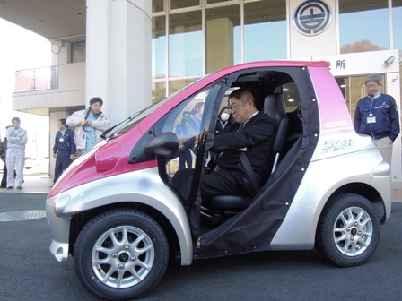 市役所玄関前で、ピンクとシルバーの三角の形をした一人乗り小型電気自動車に乗車し、ハンドルを握る市長の写真