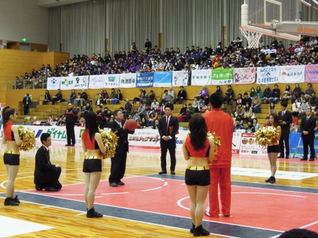 宮古市民総合体育館で大勢の観客が席に座っており、バスケットボールコート前でチアリーダーや関係者が見守る中、市長がバスケットボールをゴールに向かって投げようとしている写真