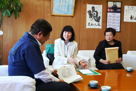 市長と童話作家の茂市久美子さんと関係者が応接室の椅子に座り、発行された新刊を市長が手に持って眺めており、茂市久美子さんが市長に話をしている様子の写真