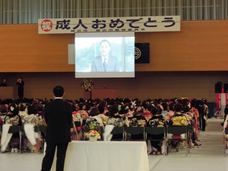 「祝成人おめでとう」と書かれた横断幕の下に俳優の渡辺謙さんが映し出されたスクリーンがあり、新成人が椅子に座り見ている写真