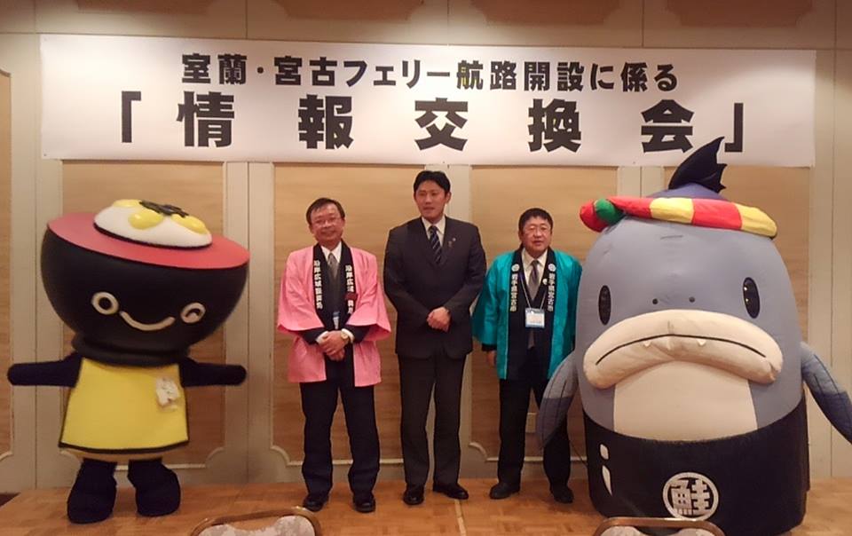 「室蘭・宮古フェリー航路開設に係る「情報交換会」」と書かれた横断幕が掛けられた壁の前で、横一列に並ぶスーツを着た男性、ピンク色の法被を着た男性、青色の法被を着た市長、わんこ兄弟、サーモンくんの記念写真