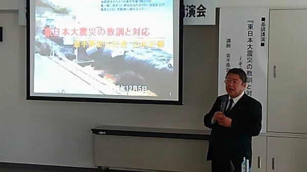 津波の映像が映されたスクリーンの横に立ち、両手でマイクを持って講演する様子の市長の写真