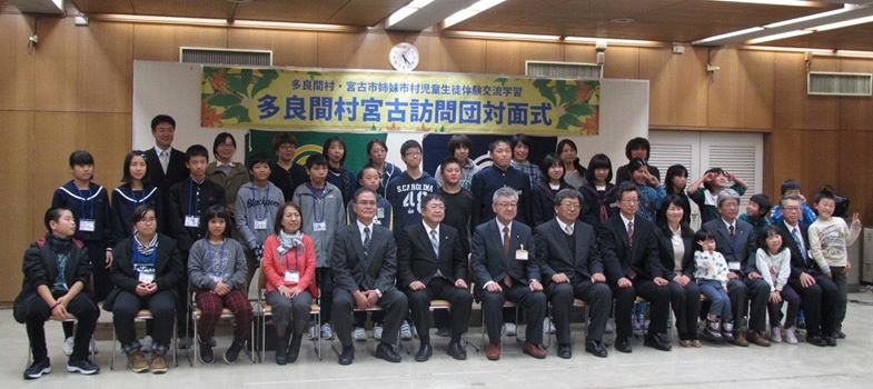 「多良間村宮古訪問対面式」と書かれた横断幕が掛けられたパネルの前で、3列に整列し記念撮影をする市長と関係者の方々、小中学生たちの写真