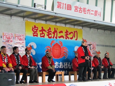 宮古毛ガニまつりと書かれ、赤いカニが描かれた大きな旗が飾られており、カニの頭飾りを付けたはっぴ姿の市長がマイクスタンドの前に立ち、話をしている写真