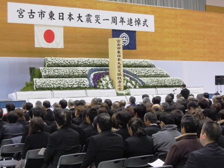 「宮古市東日本大震災一周年追悼式」と書かれた横断幕の下に国旗と宮古市章が飾られ、花祭壇が置かれた会場に多くの参加者が椅子に座っている写真