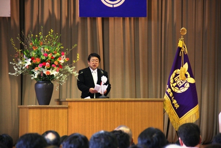 ステージ上の演壇の両隣りに生け花と校旗があり、演壇で市長が話している写真