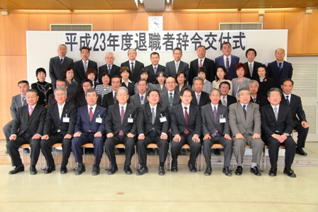 「平成23年度退職者辞令交付式」と書かれた横断幕の前でスーツを着た退職する方々が4列に並び記念撮影している写真
