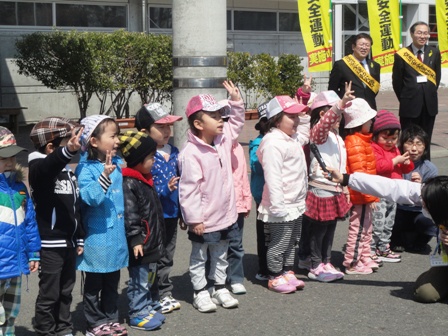 園児達が片手を上にあげて一列に並び、後ろには蛍光黄色の安全運動ののぼり旗があり、スーツを着た男性2名が見守っている写真