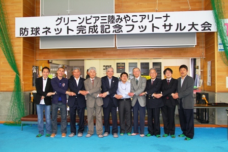 「グリーンピア三陸みやこアリーナ防球ネット完成記念フットサル大会」と書かれた横断幕の下で男性10名が手を前で交差し、交差した手を隣の人と繋ぎ記念撮影している写真