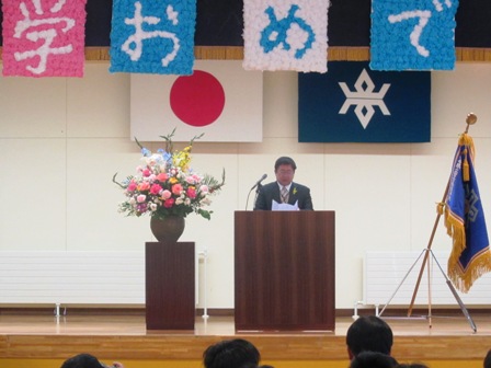 学校旗や花瓶に花が飾られた舞台で、演壇に立ち話をする市長の写真