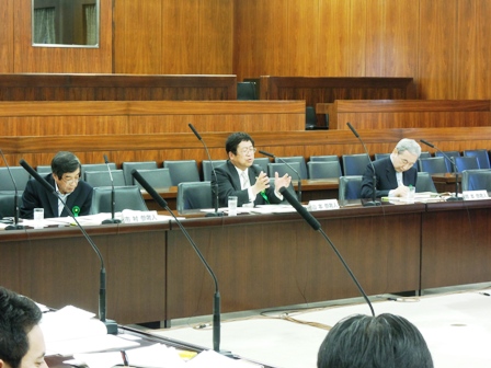 議会の席で市長が発言しており、両側に座る男性が資料を眺めている写真