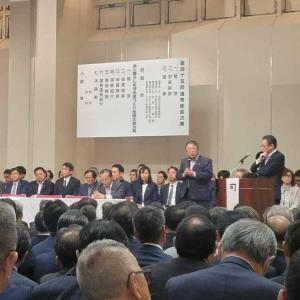 男性が立つ演台の横で、両手でマイクを持ち話をする様子の市長と、椅子に座る大勢の参加者たちの写真