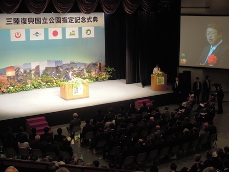 広い会場のホールで「三陸復興国立公園指定記念式典」の吊り看板があり、演壇に立つ市長がステージ右側のスクリーンに映し出されている写真