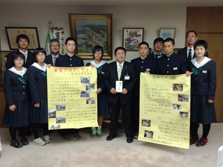 写真が貼られ文字が書き込まれた黄色い模造紙を持つ2名の男子中学生と、中心に立つ市長、周りに並ぶ中学生立ちと関係者の方々の記念写真