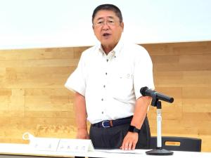 椅子の前に立ち机に置かれたスタンドマイクに向かって話をしている様子の市長の写真