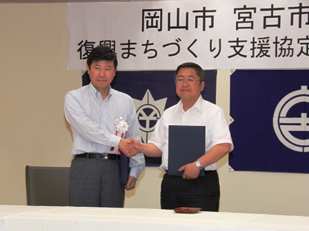壁に「岡山市と宮古市復興まちづくりの支援に関する協定」と書かれた横断幕と岡山市と宮古市の市章が飾られた前で男性と宮古市の市長が握手をし記念撮影している写真