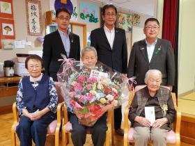 前列に椅子に腰かけている久保田イチさん、年配の女性とお祝いの花束を持った男性その後方に市長と他2人が立って記念撮影をしている写真