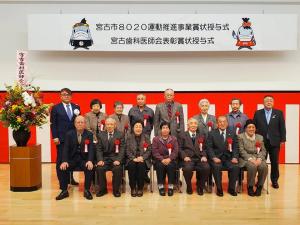 「宮古市8020運動推進事業賞状授与式 宮古歯科医師会表彰賞状授与式」と書かれた横断幕と紅白幕で飾られ、カラフルな色の豪華な壇上花が飾られたステージ上に、胸章リボンを付けた受賞者が7名前列の椅子に座り、後列に7名と市長が立っている授賞式の様子の写真