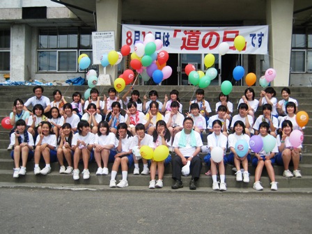 「8月10日は「道の日」です」と書かれた横断幕の前の階段に色んな色の風船を持った生徒たちと市長が3列で座り記念撮影している写真