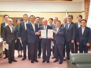 2枚の要望書を一緒に持つ市長と2名の男性、その周りに立つ1名の女性と10名の男性を写した記念写真