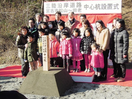 「三陸沿岸道路 宮古市松山～田老 中心杭設置式」と掲げられた横断幕の下、赤じゅうたんの上に立つ市長と小学生と関係者が中心杭の前に並んでいる写真