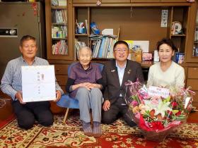 お祝いの花が置かれたご自宅で市からの記念品をご家族が持ち、市長と須藤ヱコさんと女性1人が並んで撮影した写真