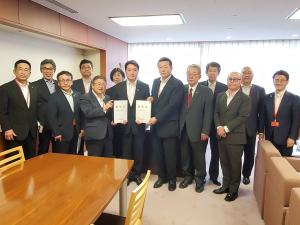 2枚の要望書を一緒に持つ市長と2名の男性、その周りに立つ1名の女性と9名の男性を写した記念写真