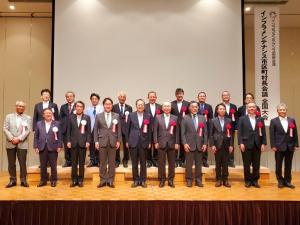 スクリーンが設置された舞台上で、胸章を着けて2列になって並ぶ市長を含む19名の関係者の方々の記念写真