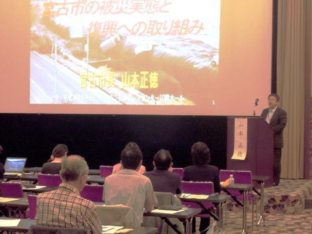 ステージのスクリーンに資料が映し出され、演壇に立って講演を行う市長と、椅子に座り講演に耳を傾けて聞く参加者の写真