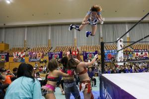 リングから場外にジャンプする女子プロレスラーと、受け止めようとする様子の3名の女子プロレスラーの写真