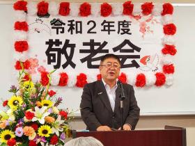 紅白の紙製の花で縁取られた令和2年度敬老会と書かれたボードをバックに 多くの花が飾ってある演台で市長があいさつをしている写真