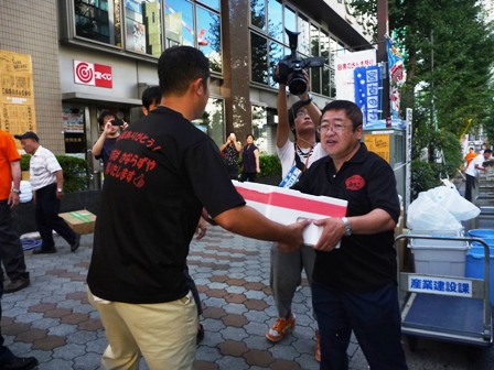 黒いTシャツを着た男性に市長が発泡スチロールを手渡しで渡しており、後ろでその様子をカメラで撮影している写真