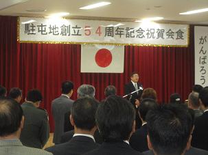 ステージ上に「駐屯地創立54周年記念祝賀会食」とかかれた横断幕の下で、 壇上に立つ市長が挨拶を行っている祝賀会の様子の写真