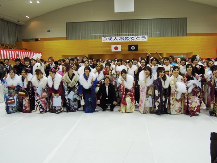 「祝成人おめでとう」と書かれた横断幕がある体育館で大勢の新成人の方々と市長が記念撮影している写真