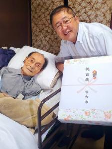 ベッドに横になる100歳を迎えた村上正吉さんと記念品を持った市長が一緒に写っている記念写真