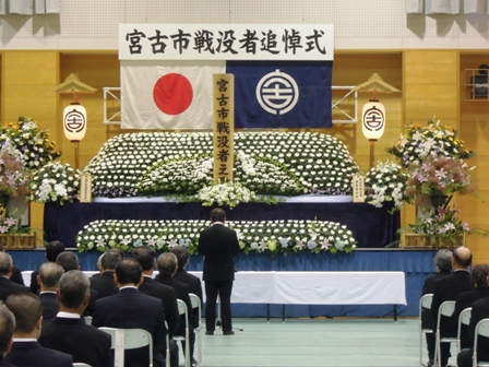 「宮古市戦没者追悼式」と書かれた横断幕の下、白い花できれいに飾られた祭壇前で市長が話をしており、その後ろに遺族の方々が椅子に座り参列している写真