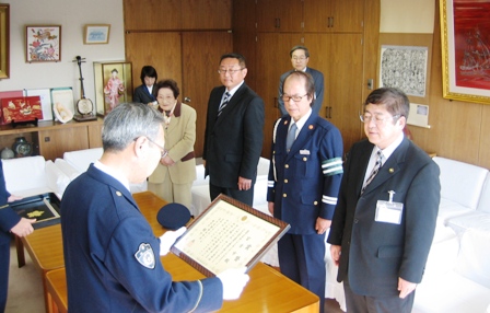 警察官の男性が額に入った称賛状を両手に持ち、向かい合って市長や関係者が横並びで立っている写真